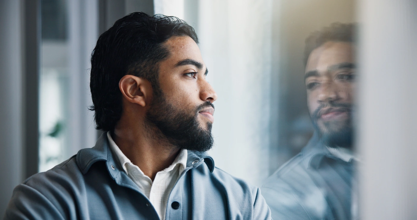 A man looking out of a window, his reflection faint in the glass